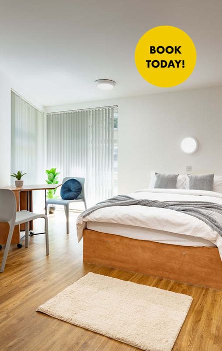 Bright, modern bedroom with a wooden floor, bed, desk, chair, plants, and a "Book Today!" label on the wall.