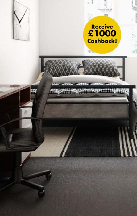 Bedroom with a neatly made bed, patterned pillows, a desk with an office chair, and a sign offering £1000 cashback.