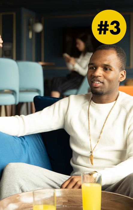 Man in a white sweater sits on a couch with a drink nearby. A person in the background is using a phone. Yellow circle with "#3" at the top.