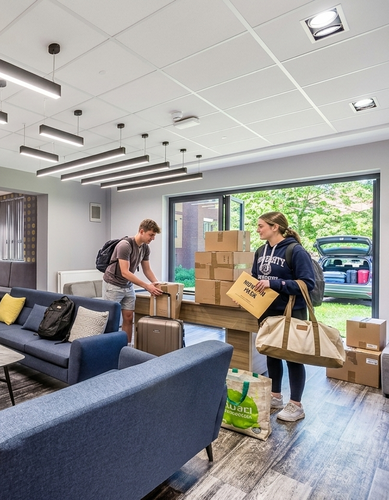 Two people in a modern living room are unpacking boxes and suitcases, with a view of a car open through a large window.