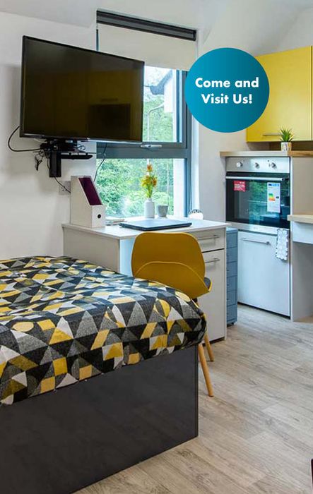 Modern studio room with a bed, desk, wall-mounted TV, and kitchen area. Text on a bubble reads "Come and Visit Us!"