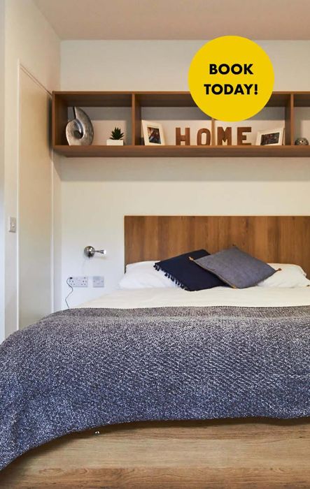 Cozy bedroom with a wooden bed, gray blanket, and decorative "HOME" letters on a shelf. A yellow "Book Today!" sign is in the corner.