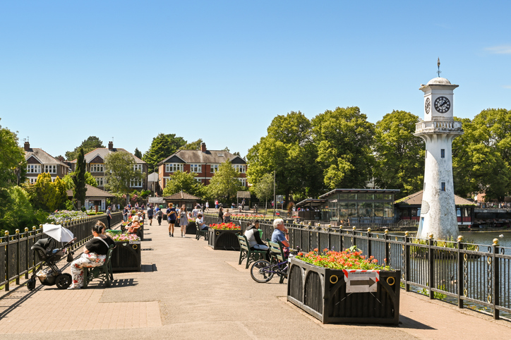 People walking and sitting on a sunny riverside path with vibrant flower planters, a clock tower, and trees in the background.
