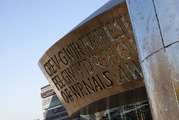 Exterior of a modern building with large, curved metallic facade featuring cut-out letters. The sky is clear and blue.