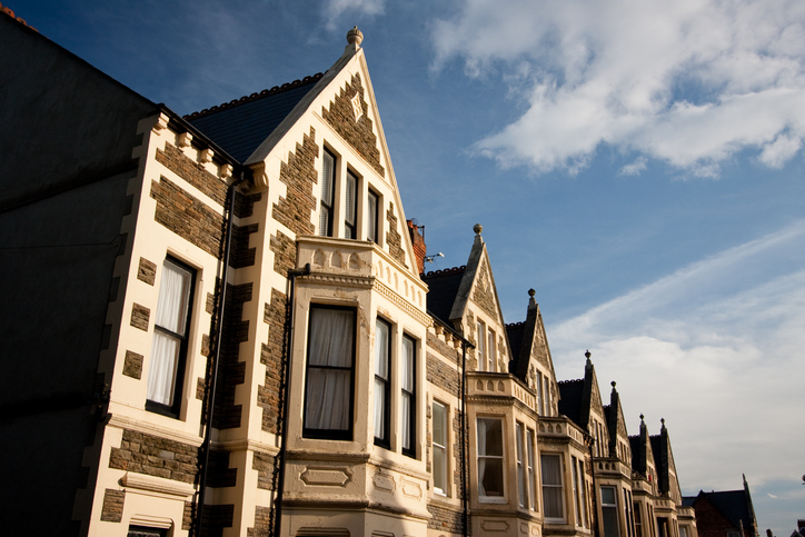 Row of Victorian-style houses with ornate gables and stone facades under a blue sky with scattered clouds.
