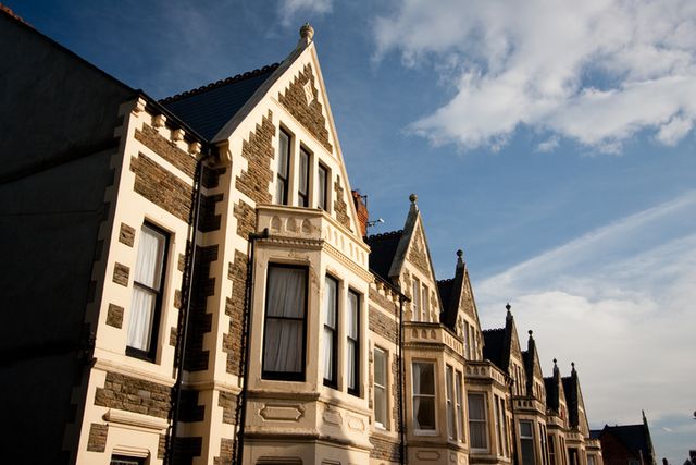 Row of Victorian-style houses with ornate gables and stone facades under a blue sky with scattered clouds.