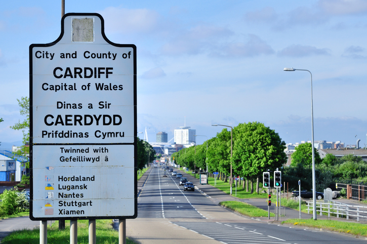 Road sign for Cardiff, Wales, with twin cities listed. Trees line the road, and buildings are visible in the distance under a cloudy sky.