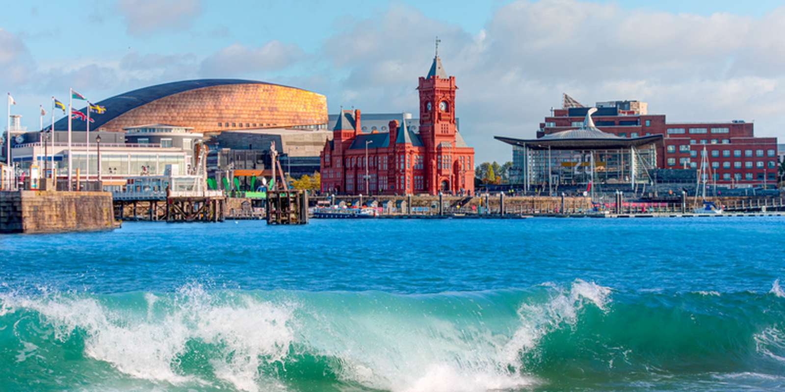 A vibrant seaside view of Cardiff Bay with the Wales Millennium Centre, Pierhead Building, and Senedd against a blue sky and crashing waves.