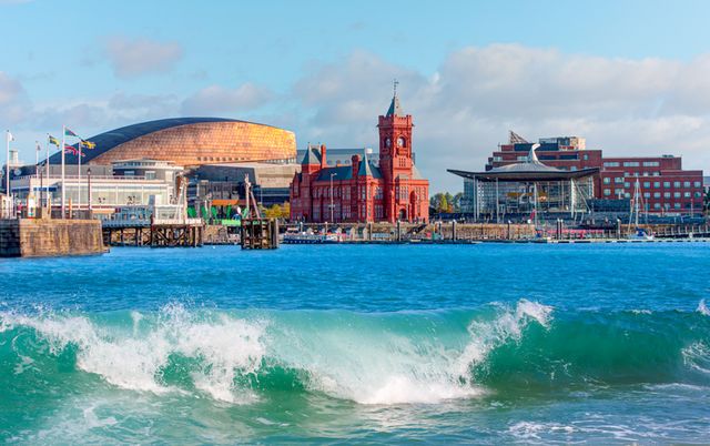 A vibrant seaside view of Cardiff Bay with the Wales Millennium Centre, Pierhead Building, and Senedd against a blue sky and crashing waves.