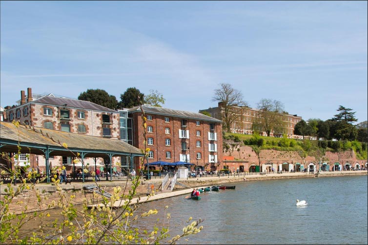Historic brick buildings along a riverside with a swan swimming, people walking, and lush greenery under a clear blue sky.