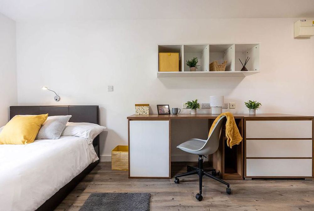 Modern bedroom with a neatly made bed, yellow accents, a wooden desk with a gray chair, and shelves holding plants and decor.
