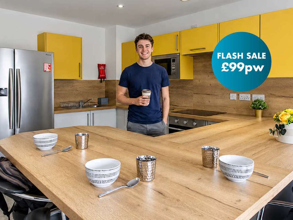 Man smiling in a modern kitchen with wooden countertops and yellow cabinets, holding a coffee. Sign reads "Flash Sale £99pw".