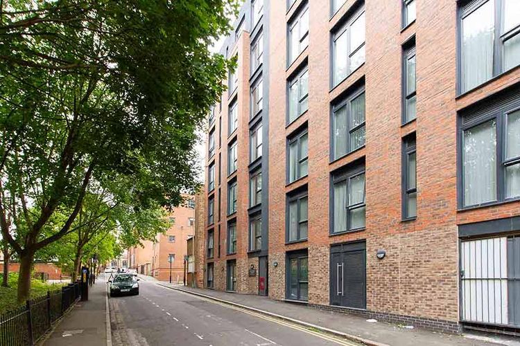 Street view of a modern brick apartment building with large windows, adjacent to a tree-lined sidewalk and parked cars.
