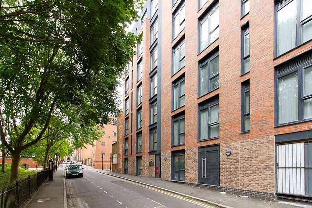 Street view of a modern brick apartment building with large windows, adjacent to a tree-lined sidewalk and parked cars.