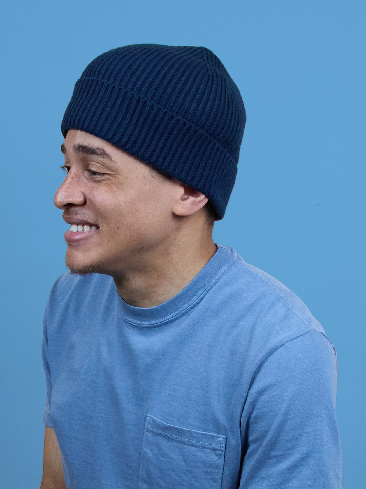 A person wearing a blue knit beanie and a blue t-shirt smiles against a solid blue background.