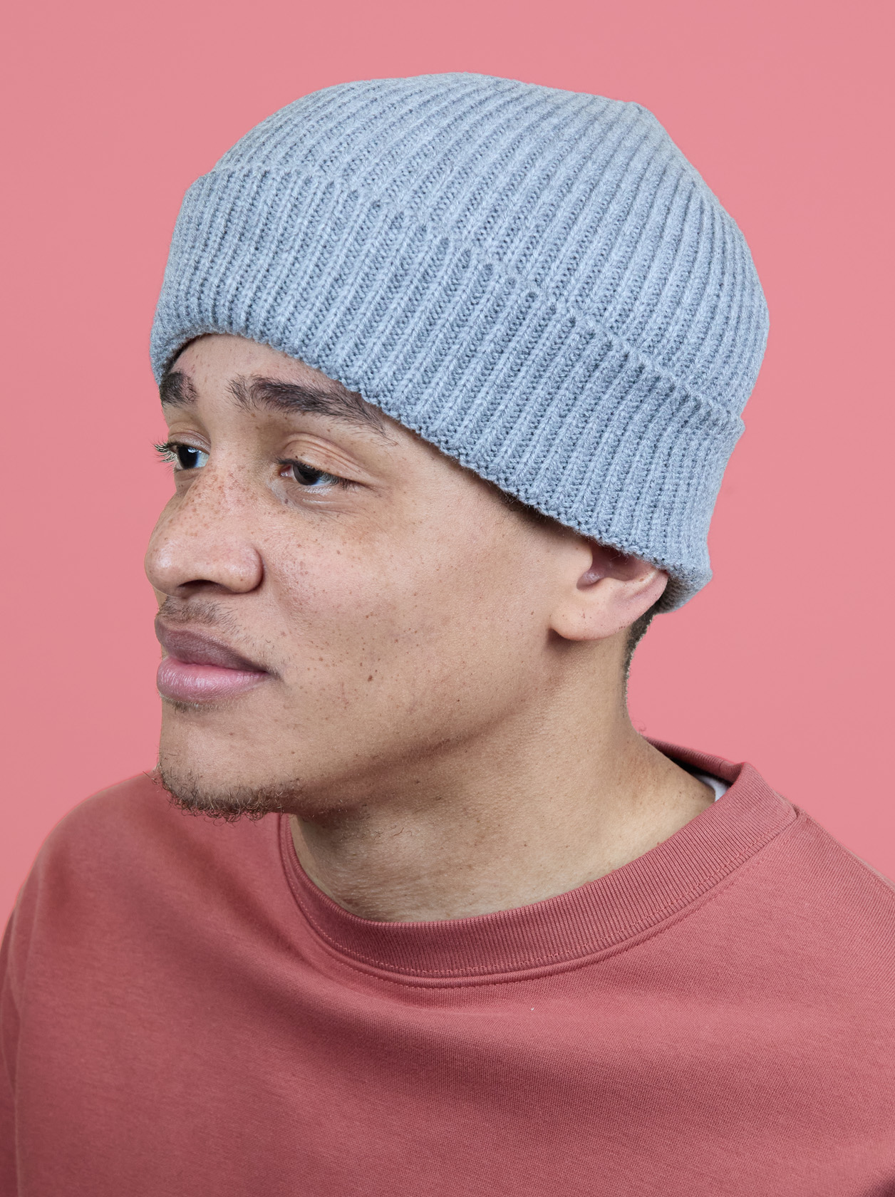 Person wearing a light blue knit beanie and a pink sweatshirt, looking to the side against a pink background.