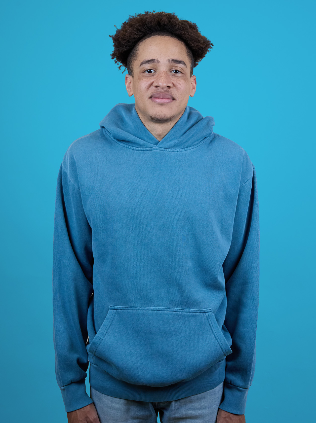 Person in a blue hoodie stands against a solid blue background, looking at the camera with a neutral expression.