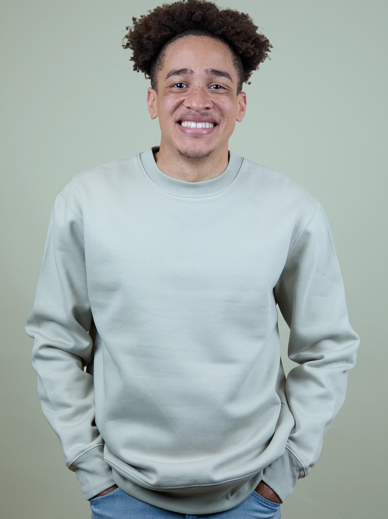 A person smiling, wearing a light gray sweatshirt and jeans, stands against a plain beige background. Hands are in pockets.