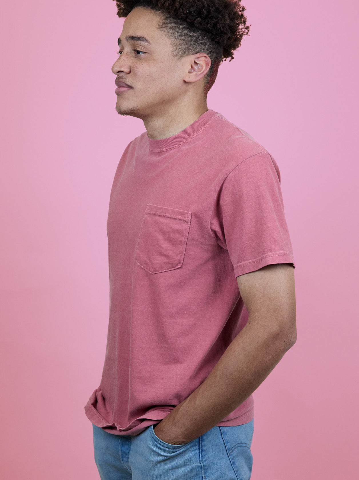 Person in a pink t-shirt and blue jeans stands against a pink background, looking to the side with hands in pockets.