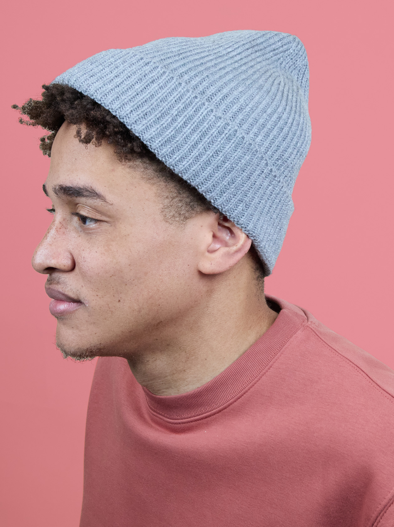 Person wearing a gray knit beanie and a pink sweatshirt, looking to the side against a pink background.