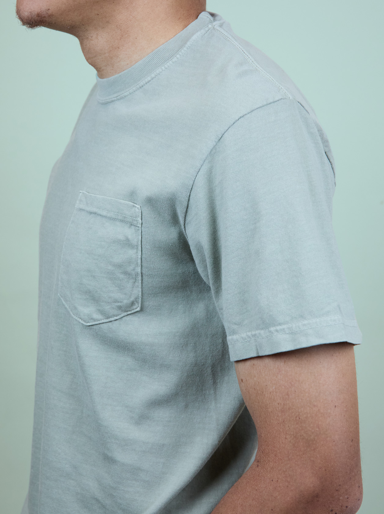 Person wearing a light gray T-shirt with a pocket, standing against a pale green background.
