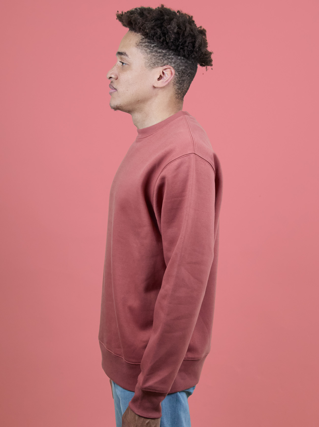 Profile of a person wearing a rust-colored sweatshirt and light blue jeans, standing against a pink background.