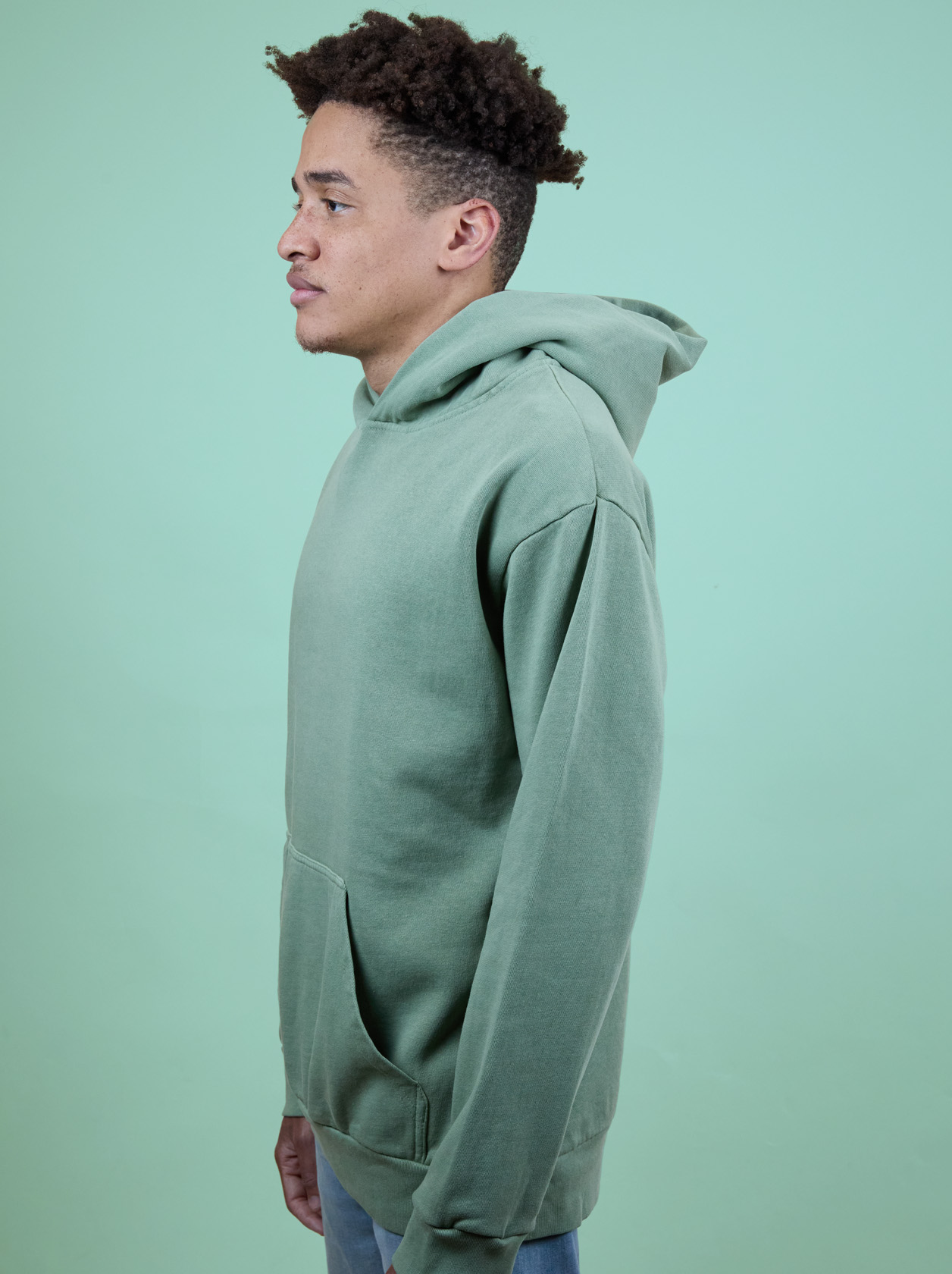 Person in a green hoodie stands sideways against a light green background, hands in pockets, looking forward.