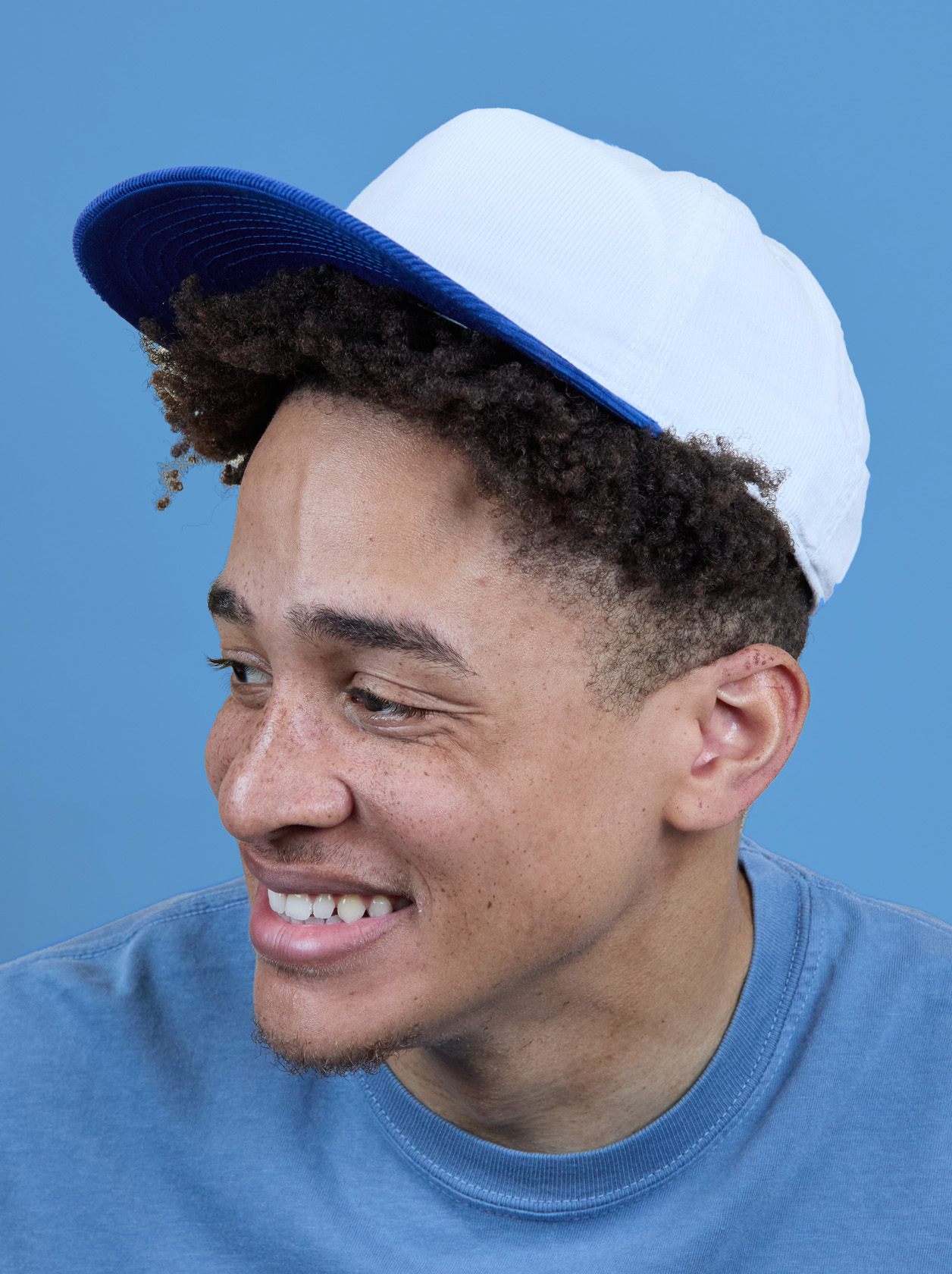 Smiling person wearing a white and blue cap and a blue shirt, looking to the side against a solid blue background.