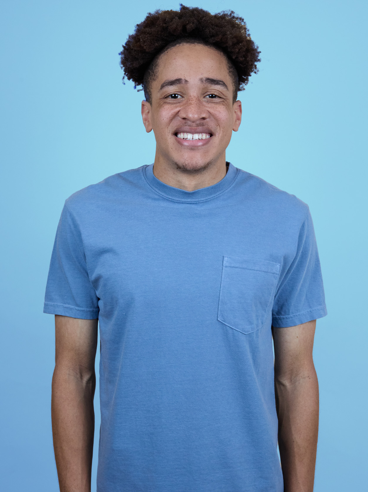 Person with curly hair smiling, wearing a blue T-shirt, standing against a light blue background.