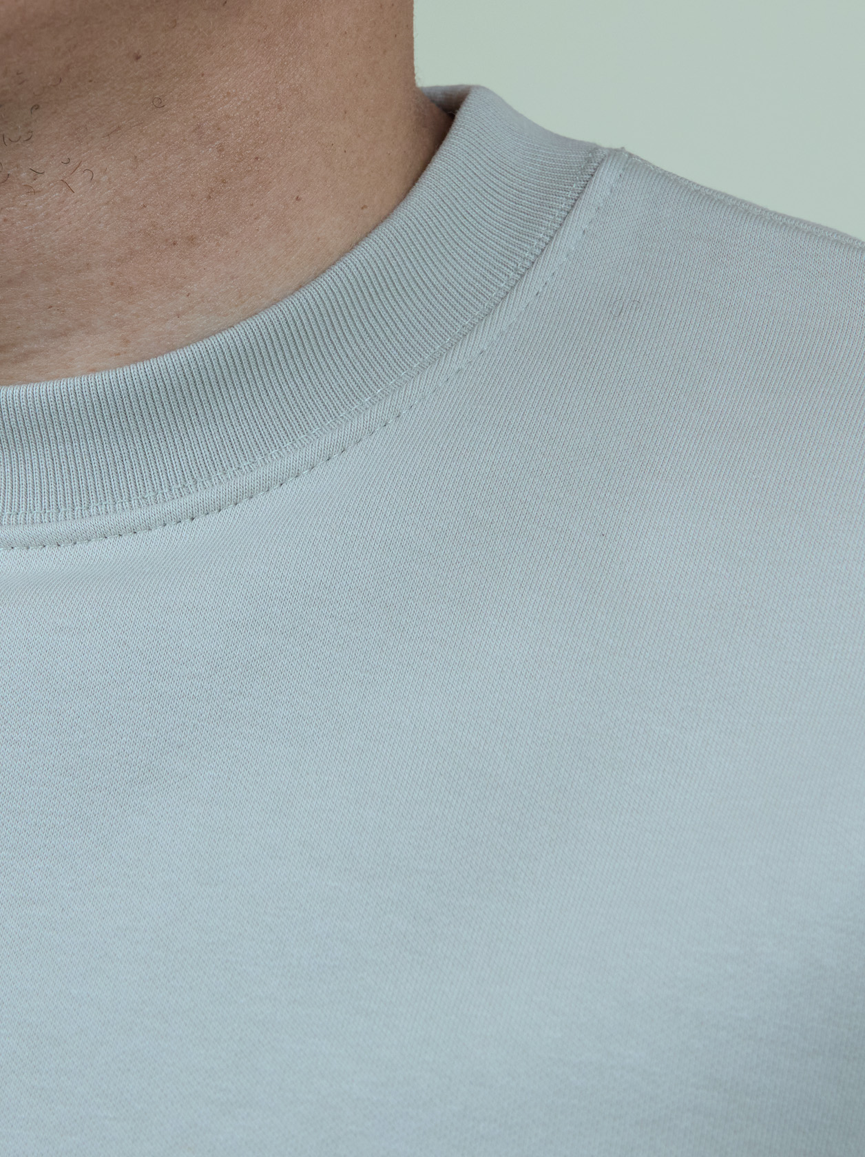 Close-up of a person wearing a light gray crew neck sweatshirt against a light background, showing part of the neck and shoulder.