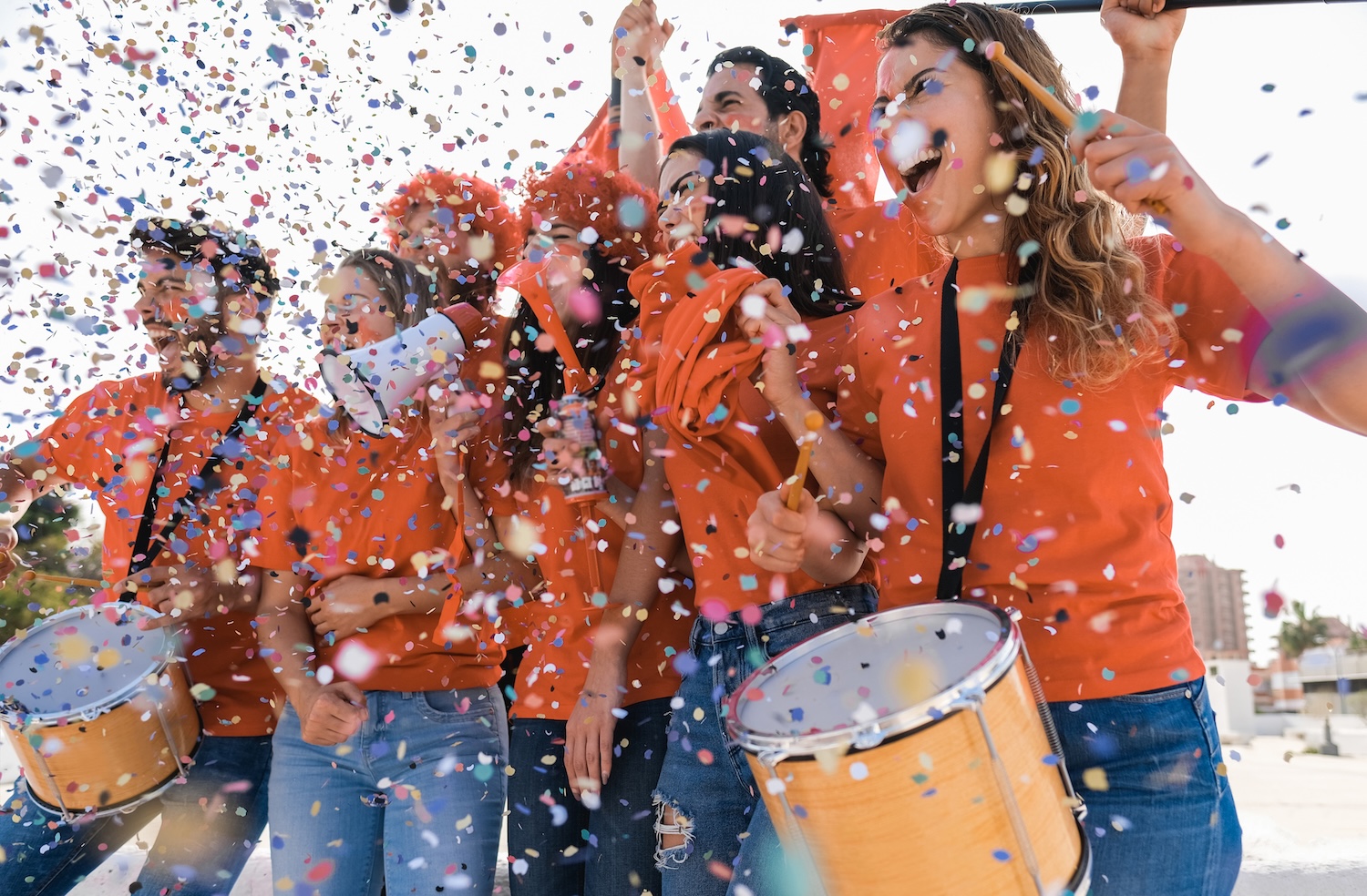 Group of people in orange shirts celebrating with confetti, holding drums and smiling outdoors.