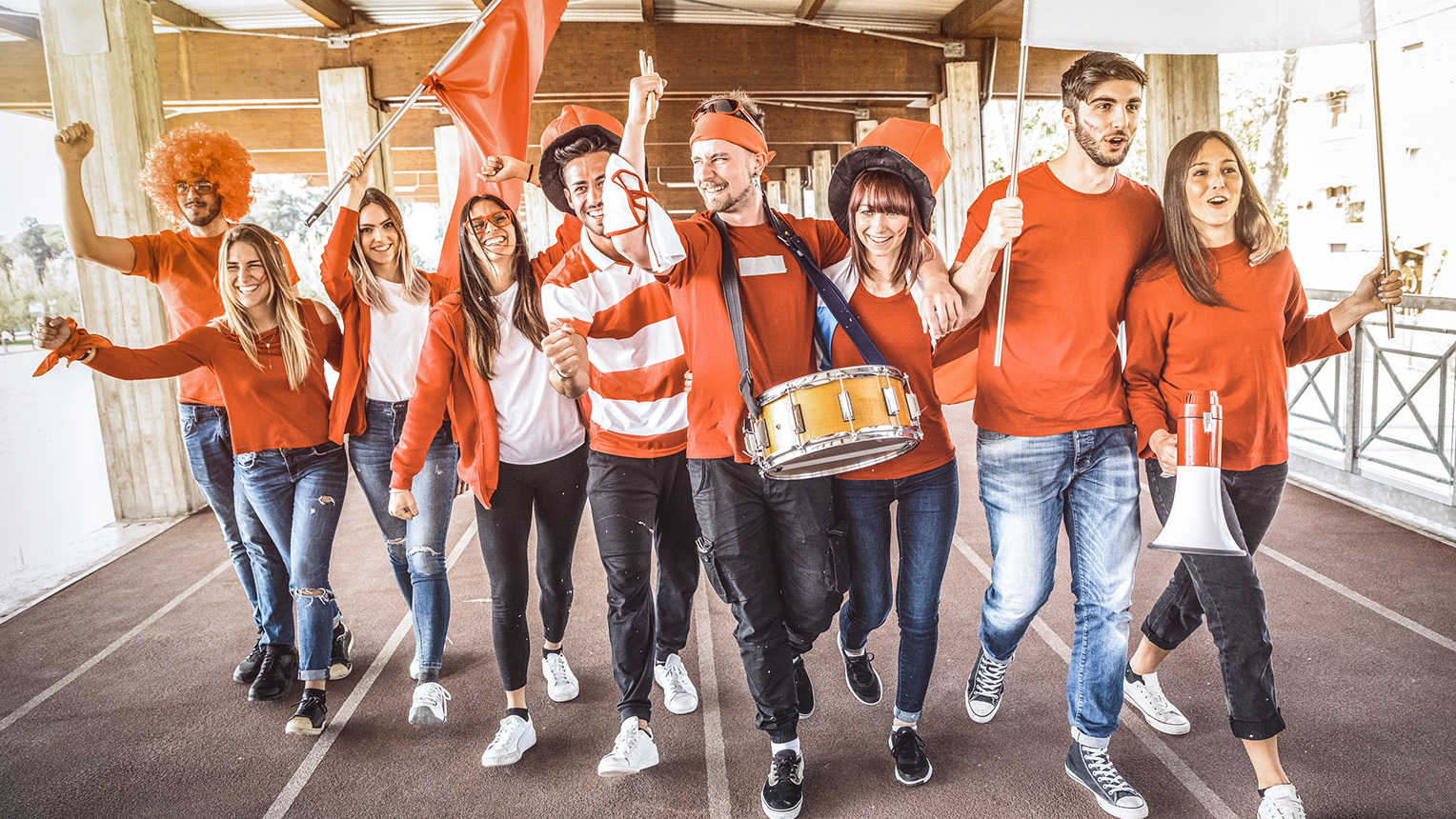 A group of enthusiastic people in red clothing, holding flags and a drum, cheering and walking together indoors.