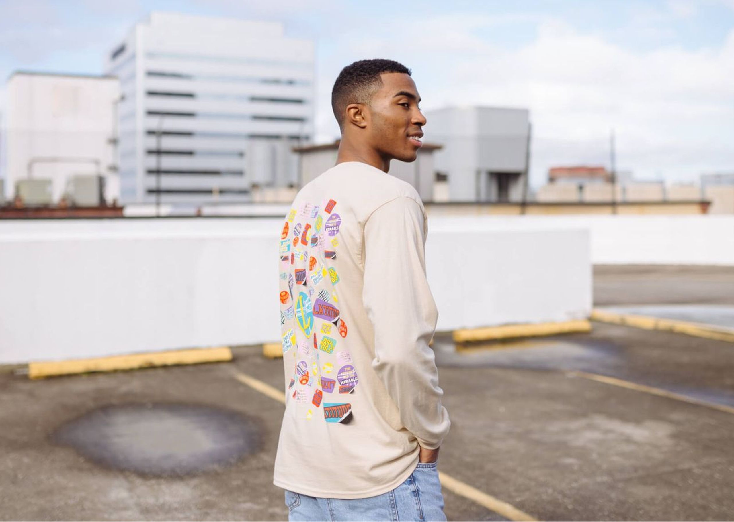 Man in a beige long-sleeve shirt with colorful patterns on the back, standing in an empty rooftop parking lot, looking over his shoulder.
