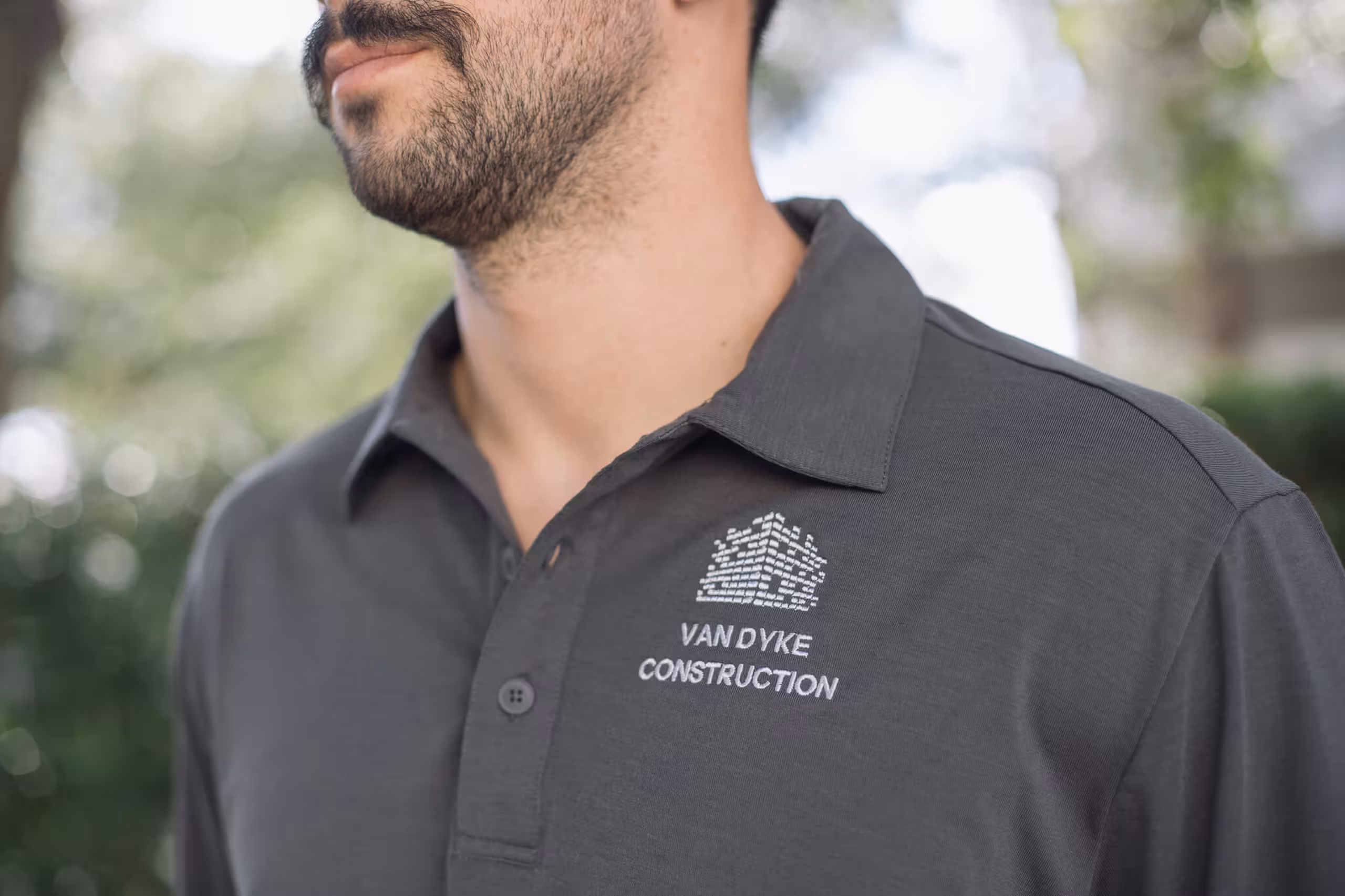 A man with a mustache wears a polo with a construction company's logo embroidered on it 