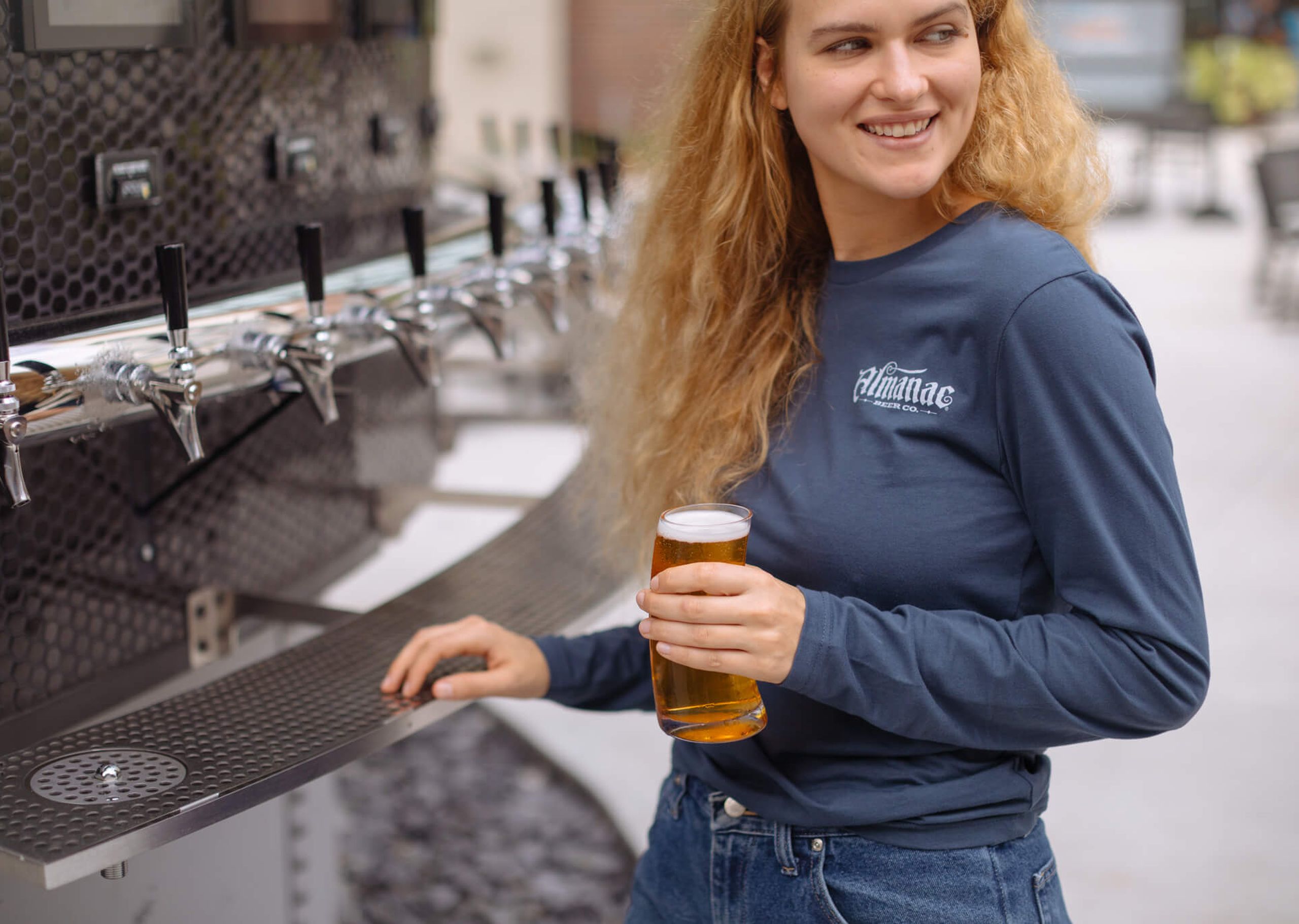 A girl drinks a glass of beer while wearing a long sleeved custom t-shirt with Almanac Beer Co. printed on it 