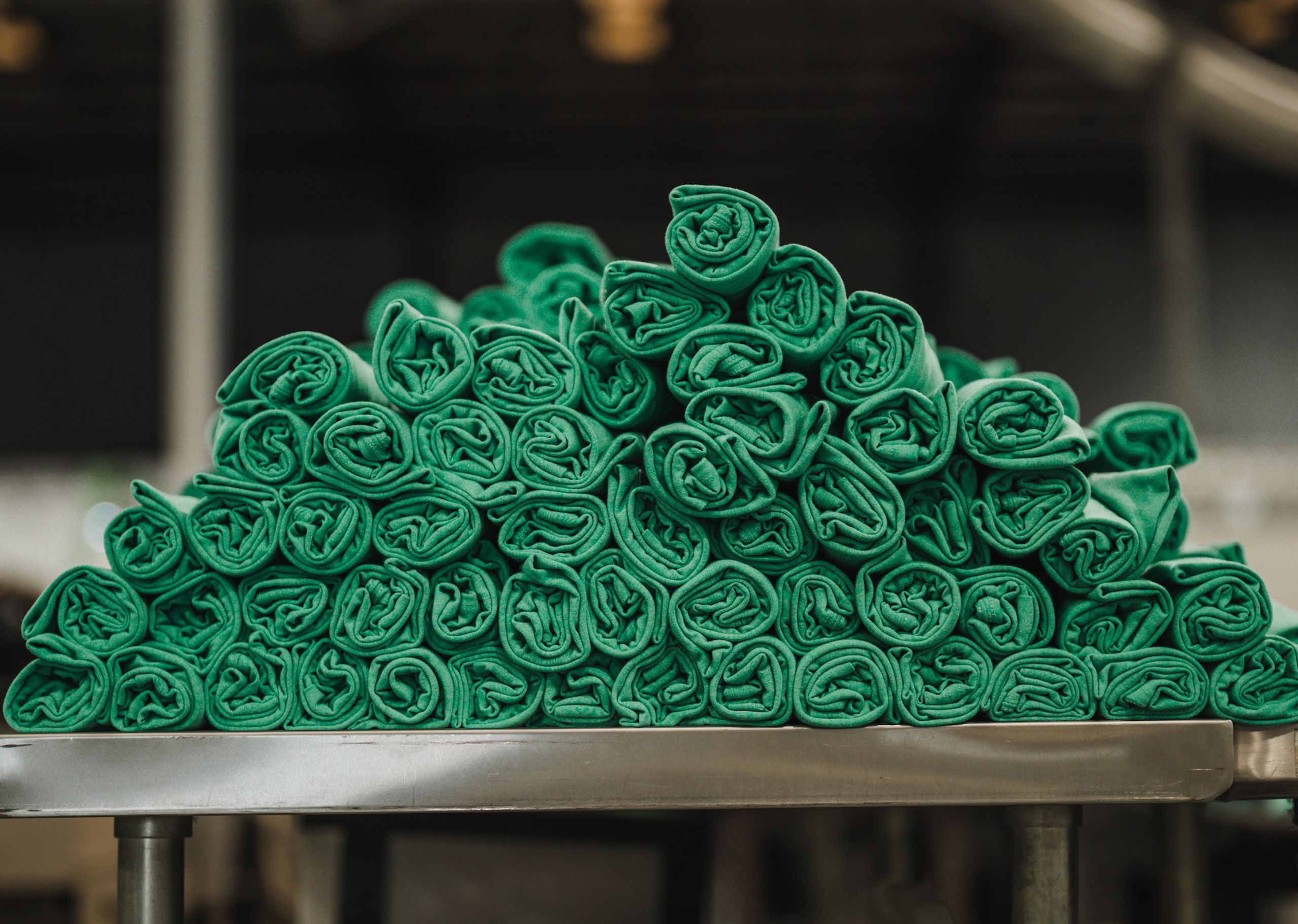 A stack of neatly rolled green towels on a metal table, set against a blurred industrial background.