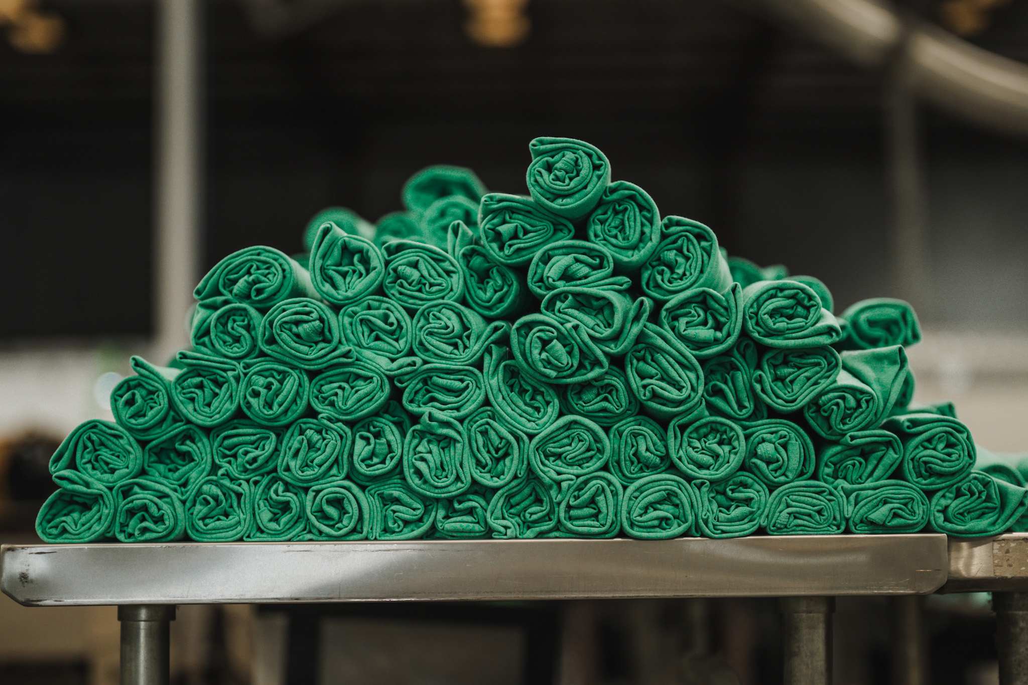 A stack of neatly rolled green towels on a metal table, set against a blurred industrial background.
