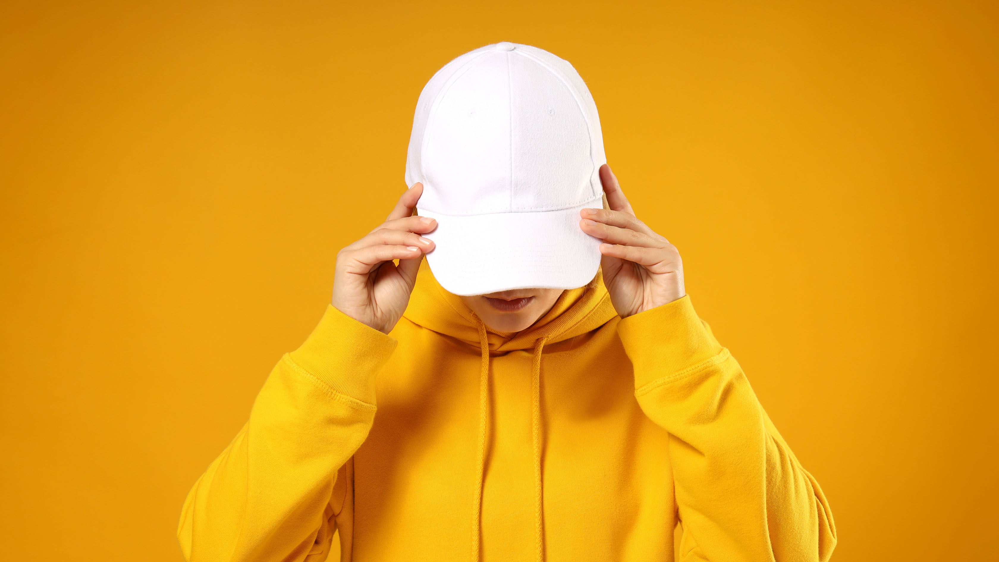 Person in a yellow hoodie adjusting a white baseball cap, set against a solid orange background.