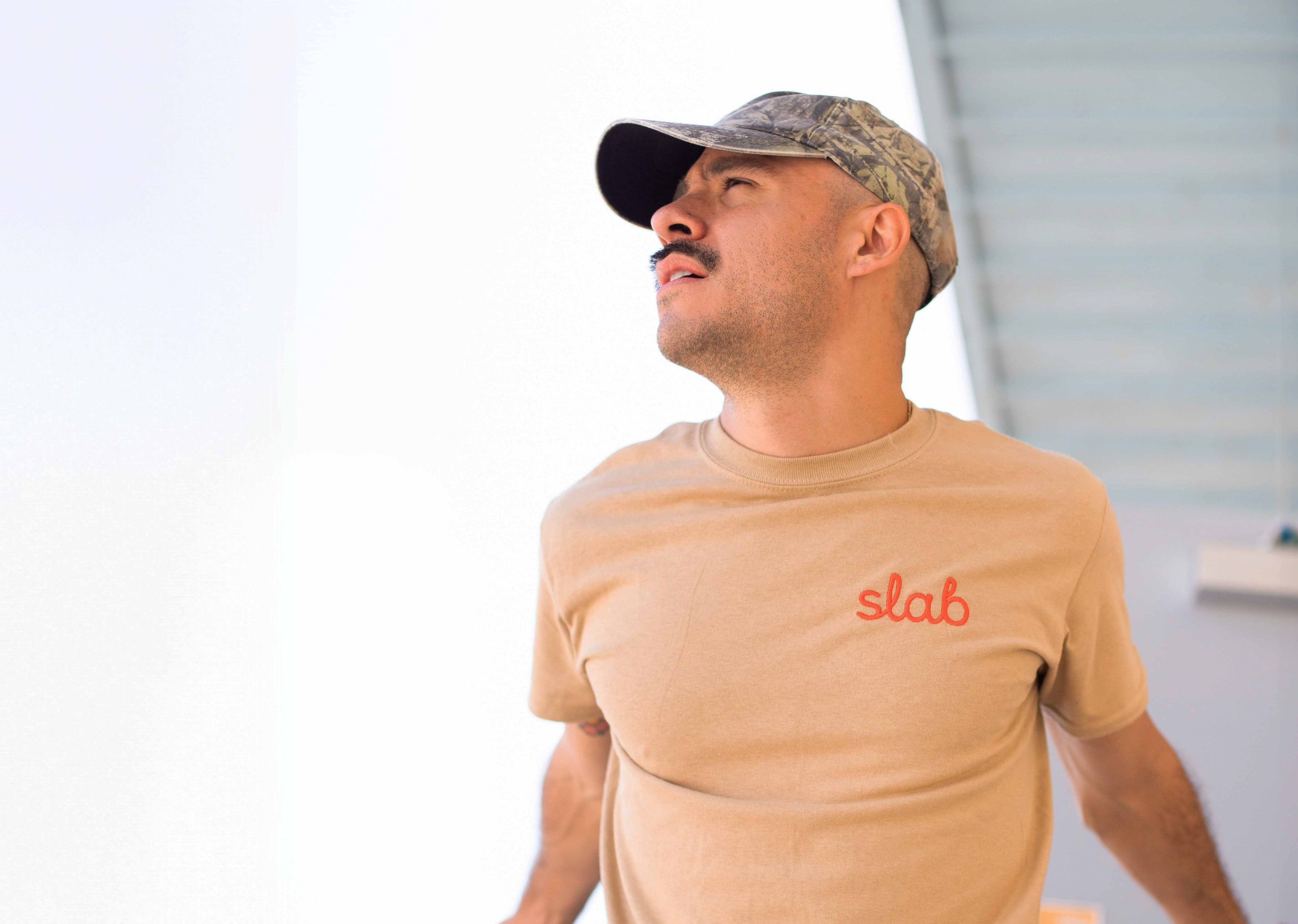 man wearing a tan t-shirt with slab embroidered on the top right in orange