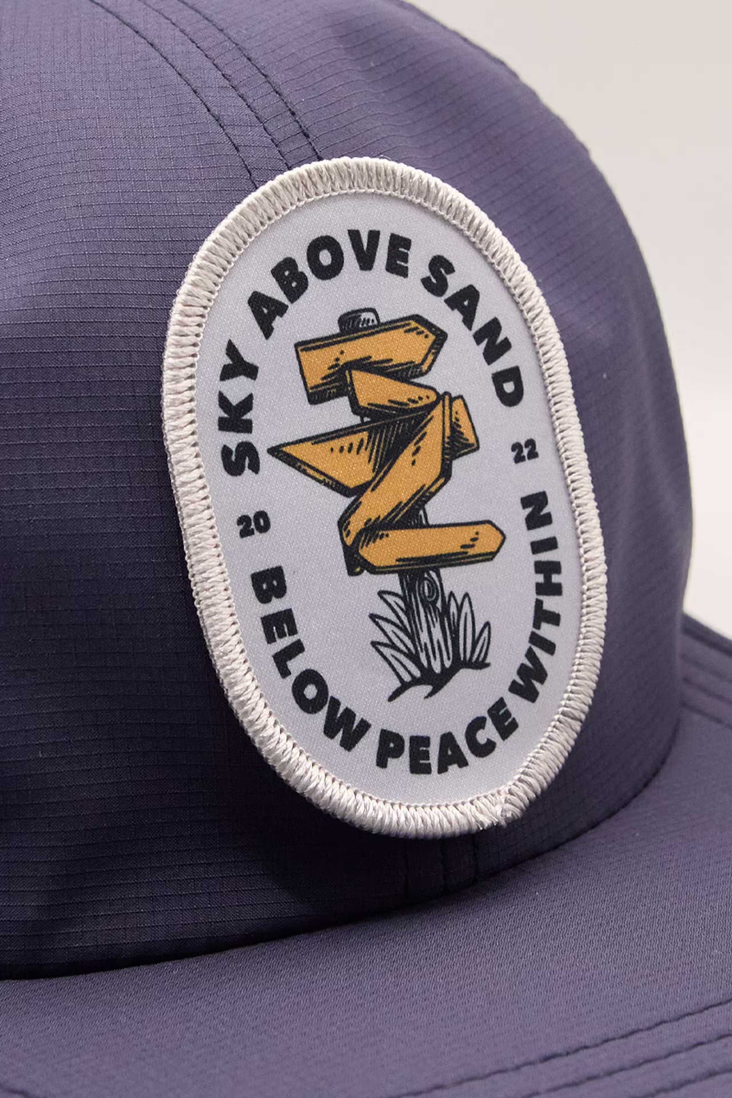 navy cap with a circular patch featuring a wooden signpost design and the text "Sky Above Sand Below Peace Within."
