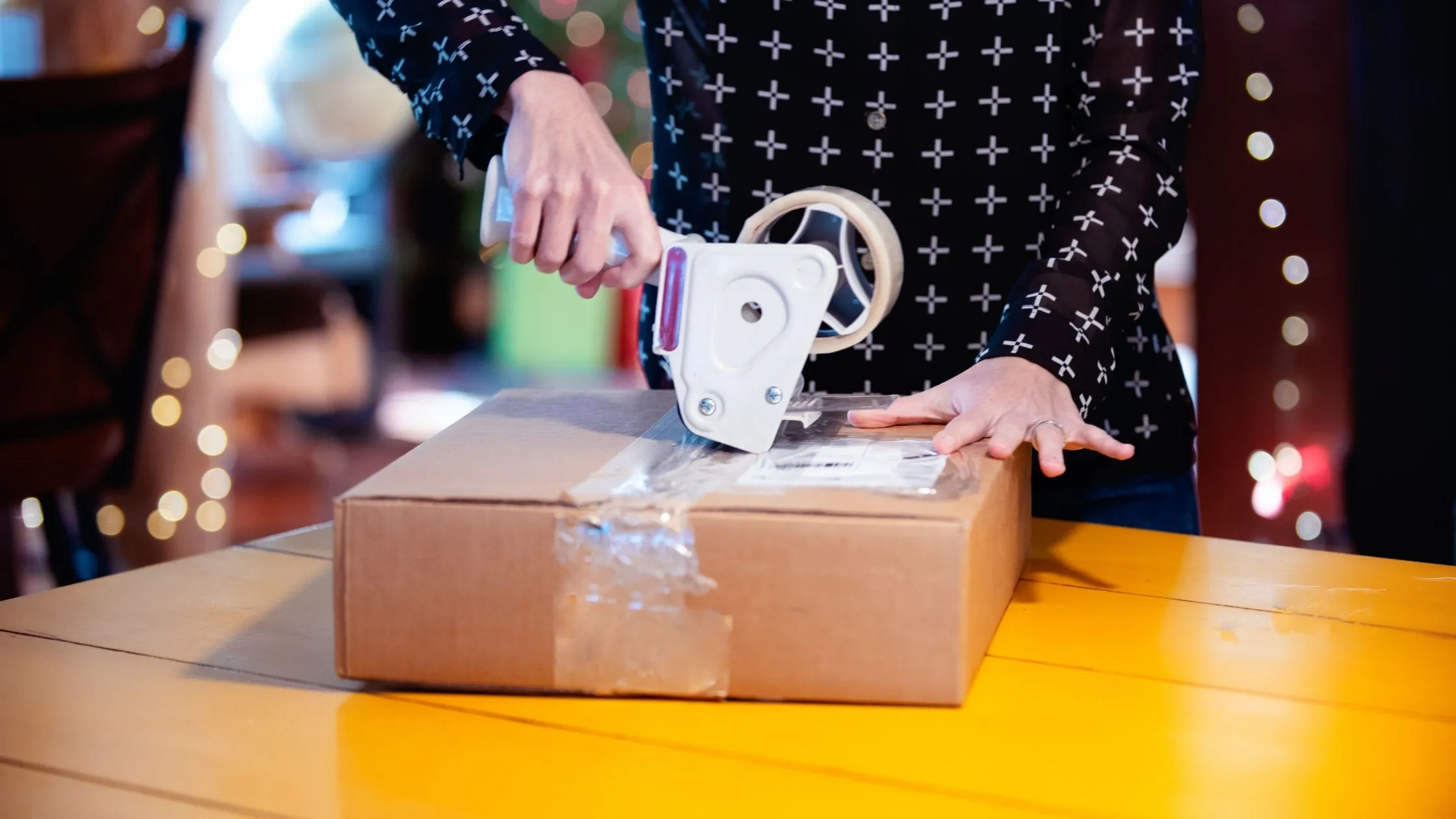 Person sealing a cardboard box with packing tape on a yellow table, surrounded by blurred lights in the background.