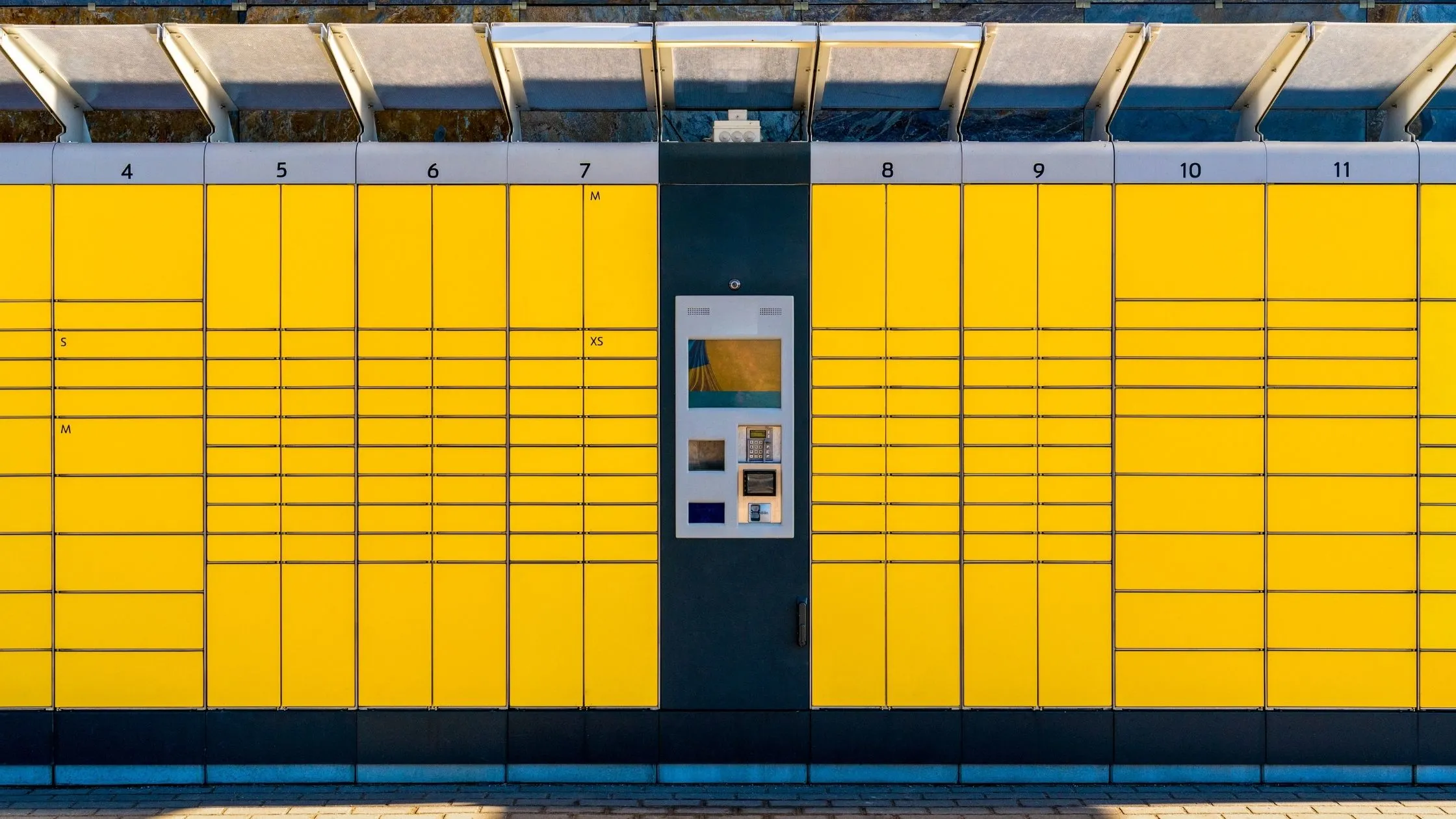 Yellow parcel locker units with numbered doors and a central control panel, set outdoors under a clear sky.