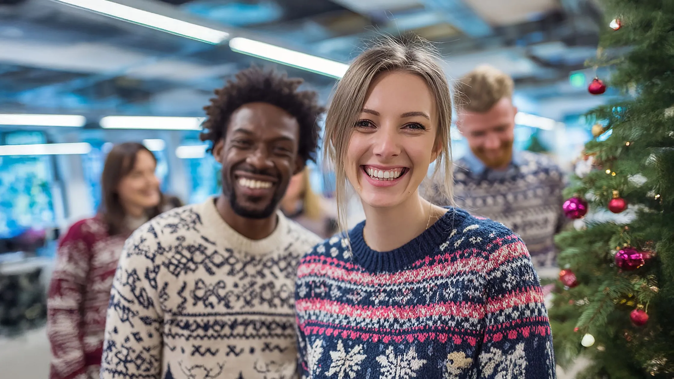 A team smiling in a festive setting, wearing patterned sweaters near a decorated Christmas tree with blurred lights in the background.