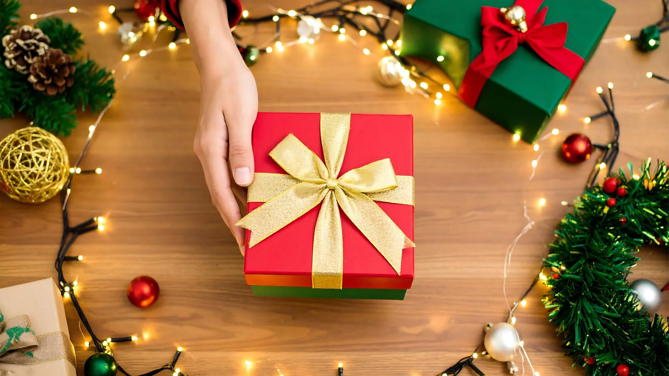 A hand reaching for a red gift box with a gold ribbon, surrounded by festive lights, green garland, and ornaments on a wooden table.