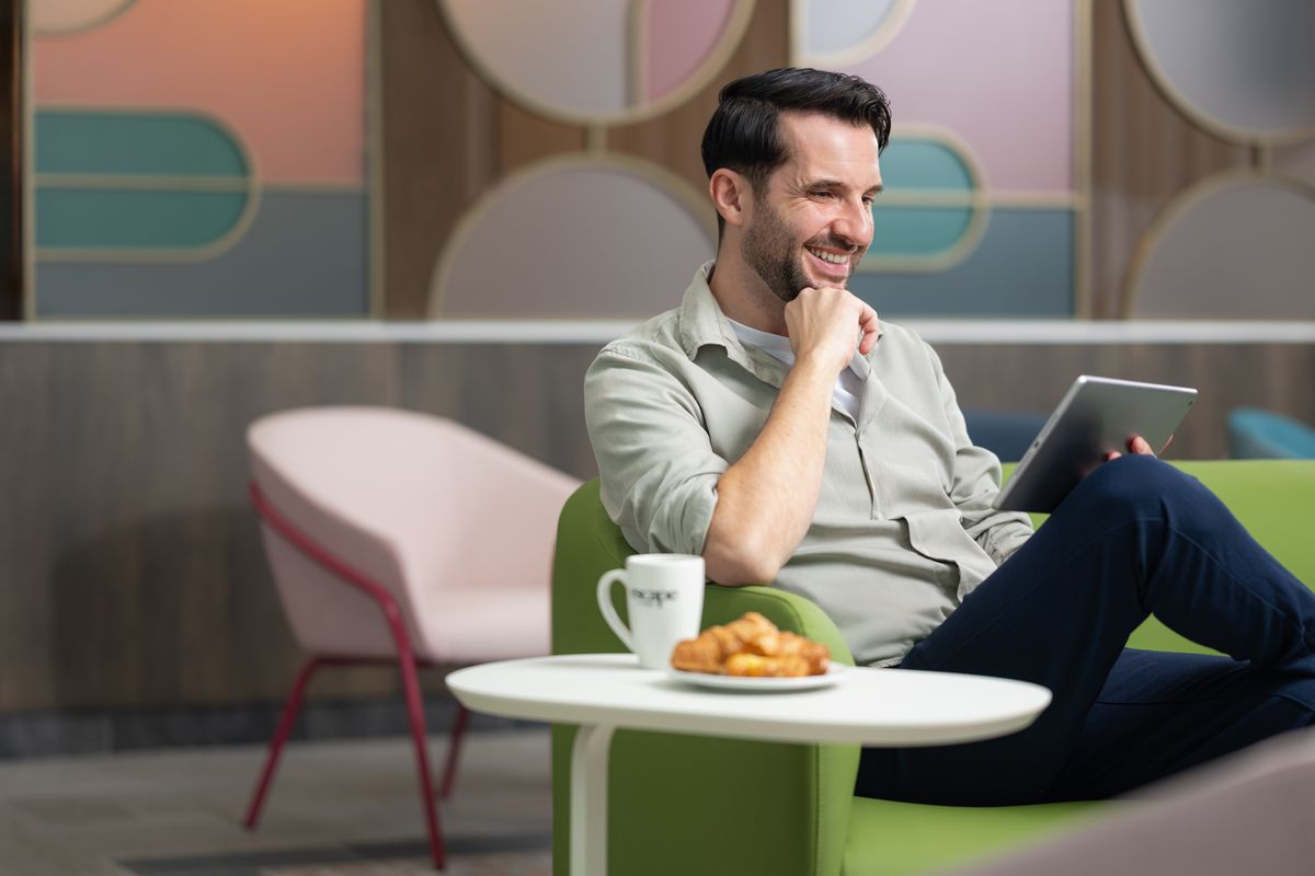 Man smiling while sitting on a green chair, holding a tablet, with a coffee mug and croissant on a table in a modern lounge.