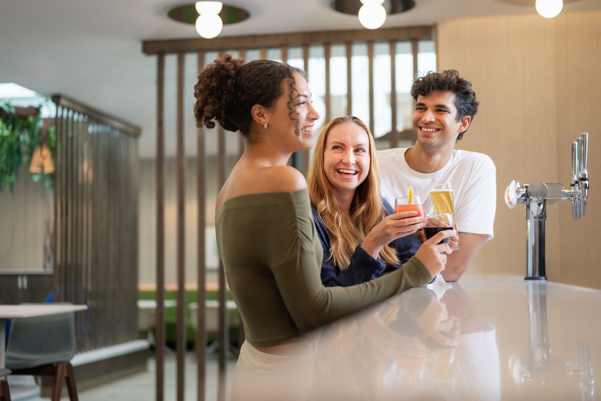 Three people standing at a bar, smiling and holding drinks, with soft lighting and modern decor in the background.