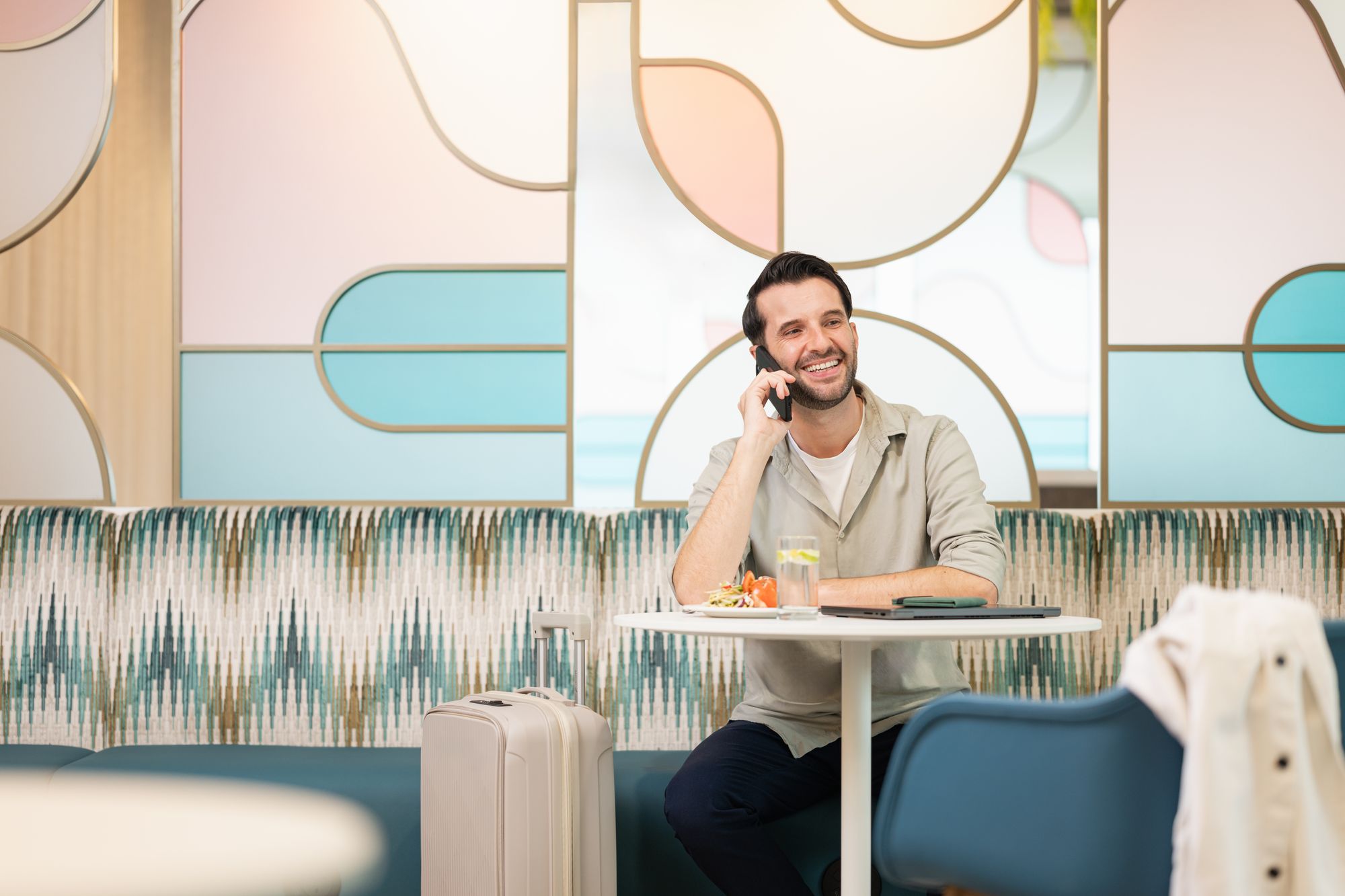 A man sitting at a table with a suitcase, talking on the phone, smiling, with a drink and food in front of him, in a modern, colorful lounge.