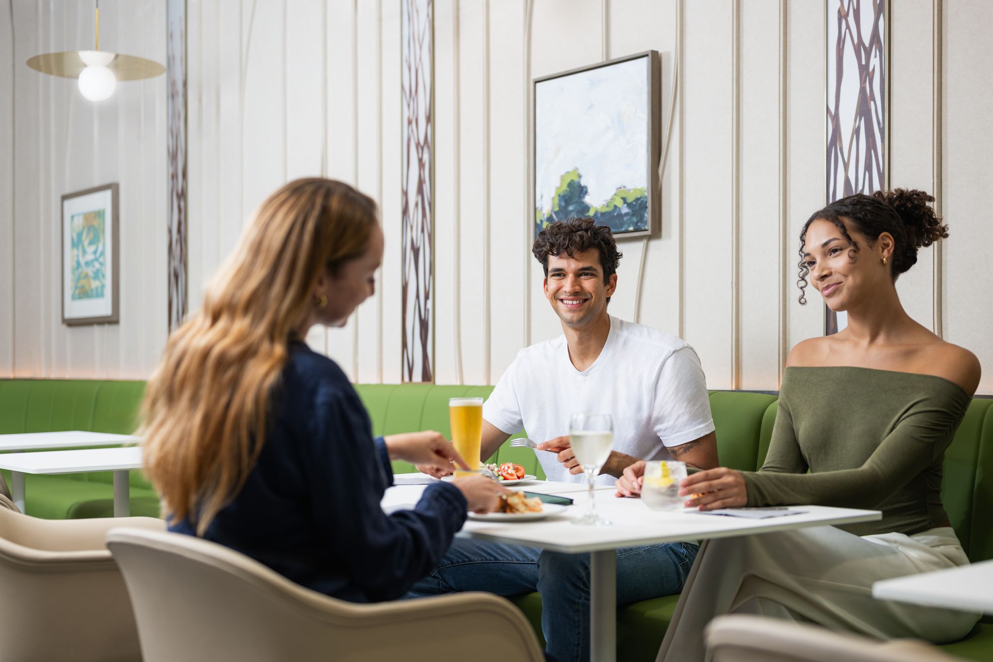 Three people sitting at a table in a bright lounge, smiling and chatting while enjoying drinks and food.