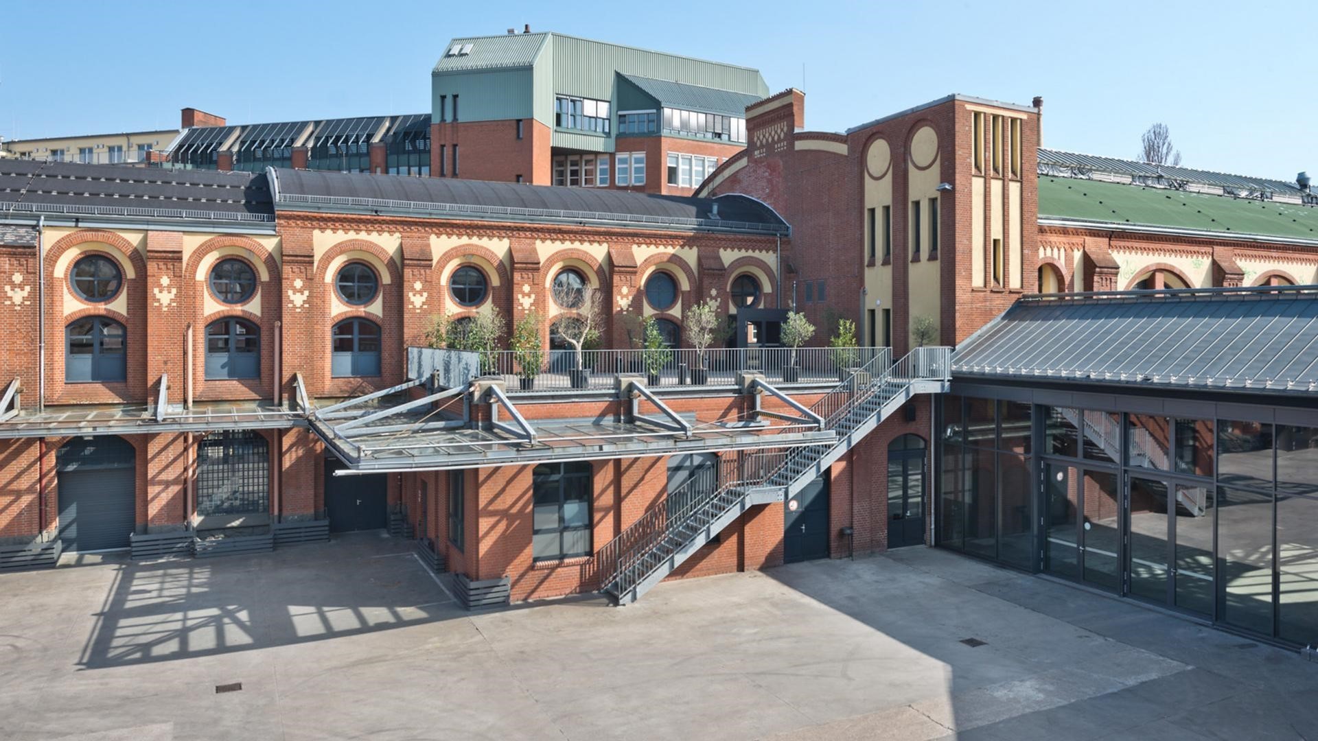 An aerial view of a large building with a staircase and a glass roof. The building has many windows and doors, and it is surrounded by other buildings. There are potted plants on the glass roof, and the building has a unique design with a combination of brick and glass walls. The building is located in an open area with a clear sky.