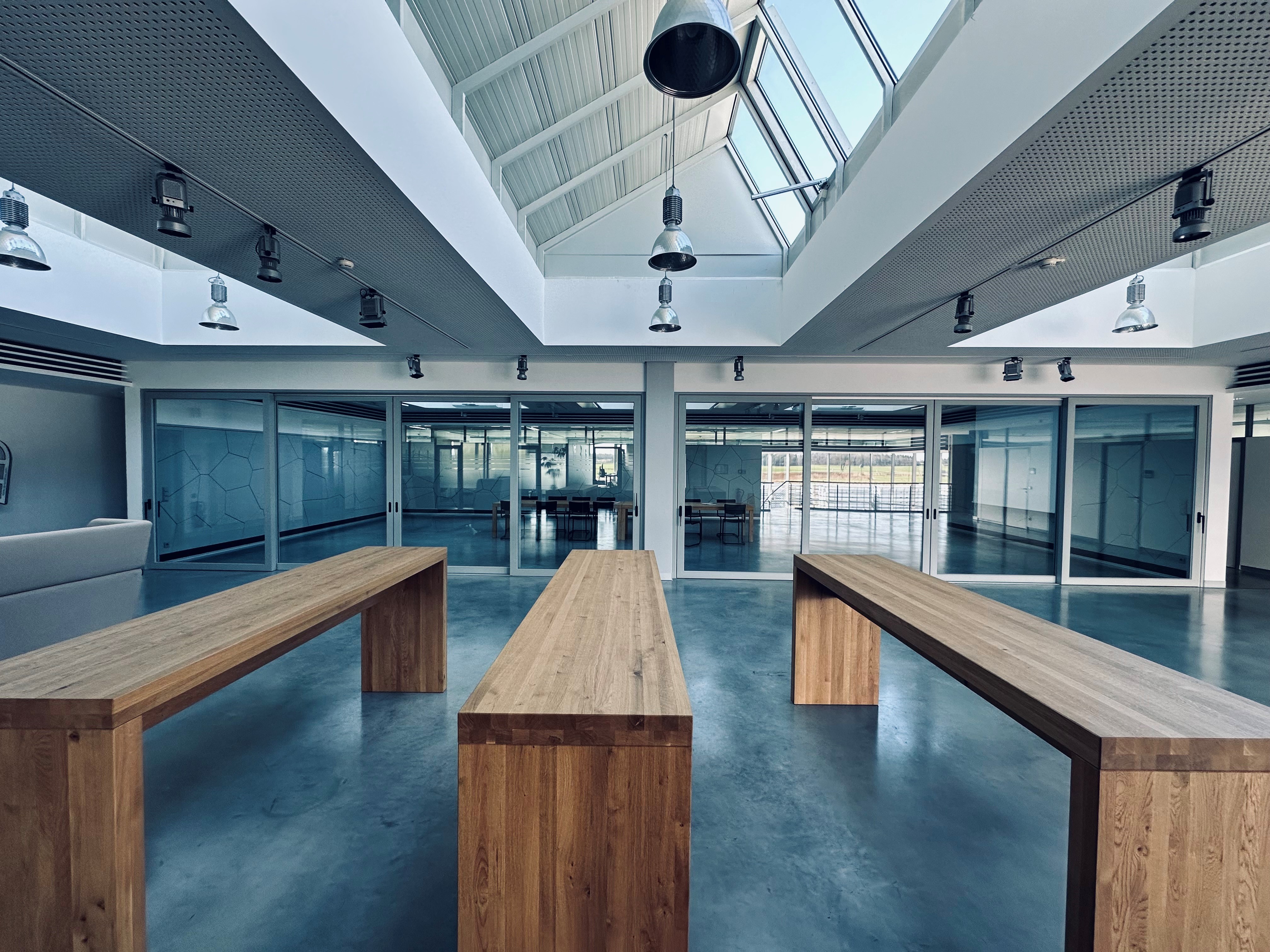 Three wooden benches in a modern office space with glass walls and a skylight, illuminated by hanging lights.